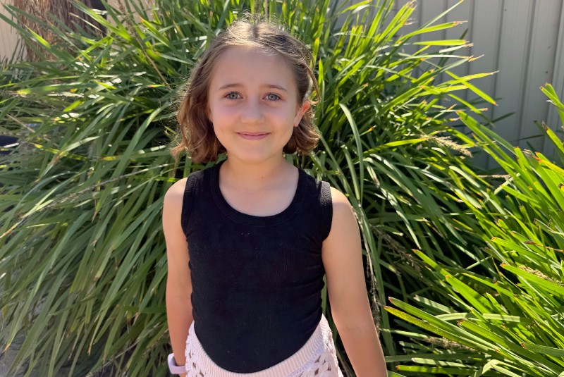 A primary-school aged girl smiles to the camera. She has short, light brown hair and stands in front of a bright green plant.